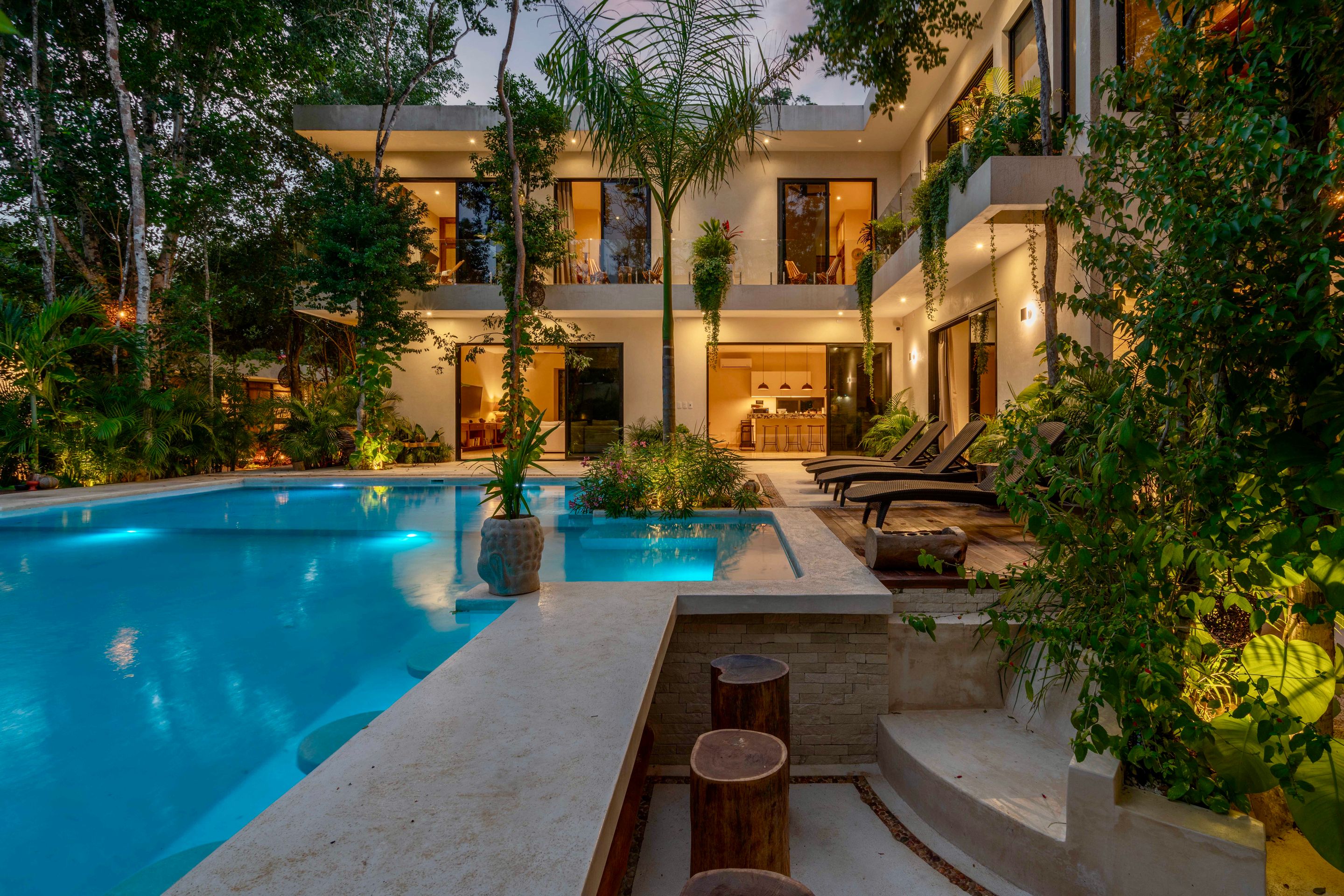 A lit swimming pool at dusk beside a warmly glowing house